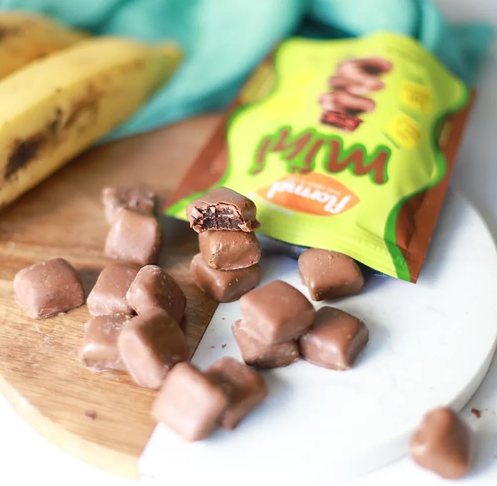 Close-up da embalagem sobre superfície de madeira, com tábua de mármore branca, pano azul esverdeado e uma banana ao fundo.