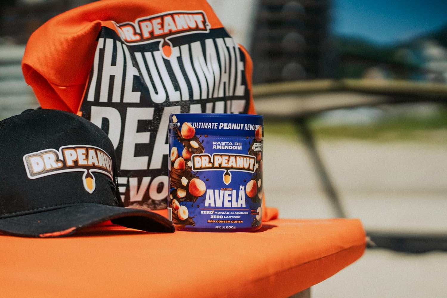 Pote de creme de avelã em perspectiva frontal com fundo desfocado em tons de azul, verde e cinza, sugerindo ambiente ao ar livre.