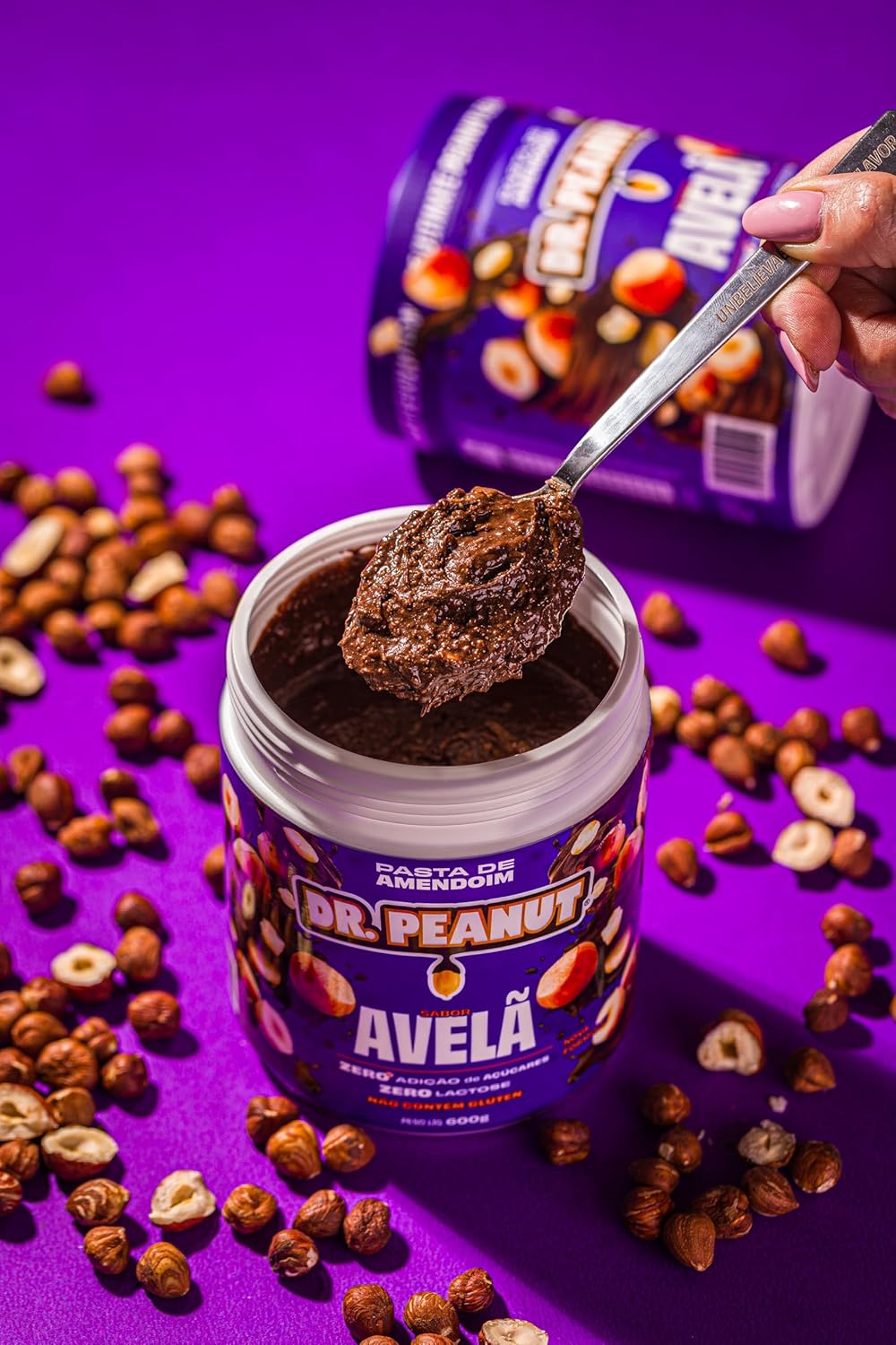 Close-up de pote de creme de avelã com ingredientes ao redor, sobre fundo liso e roxo.